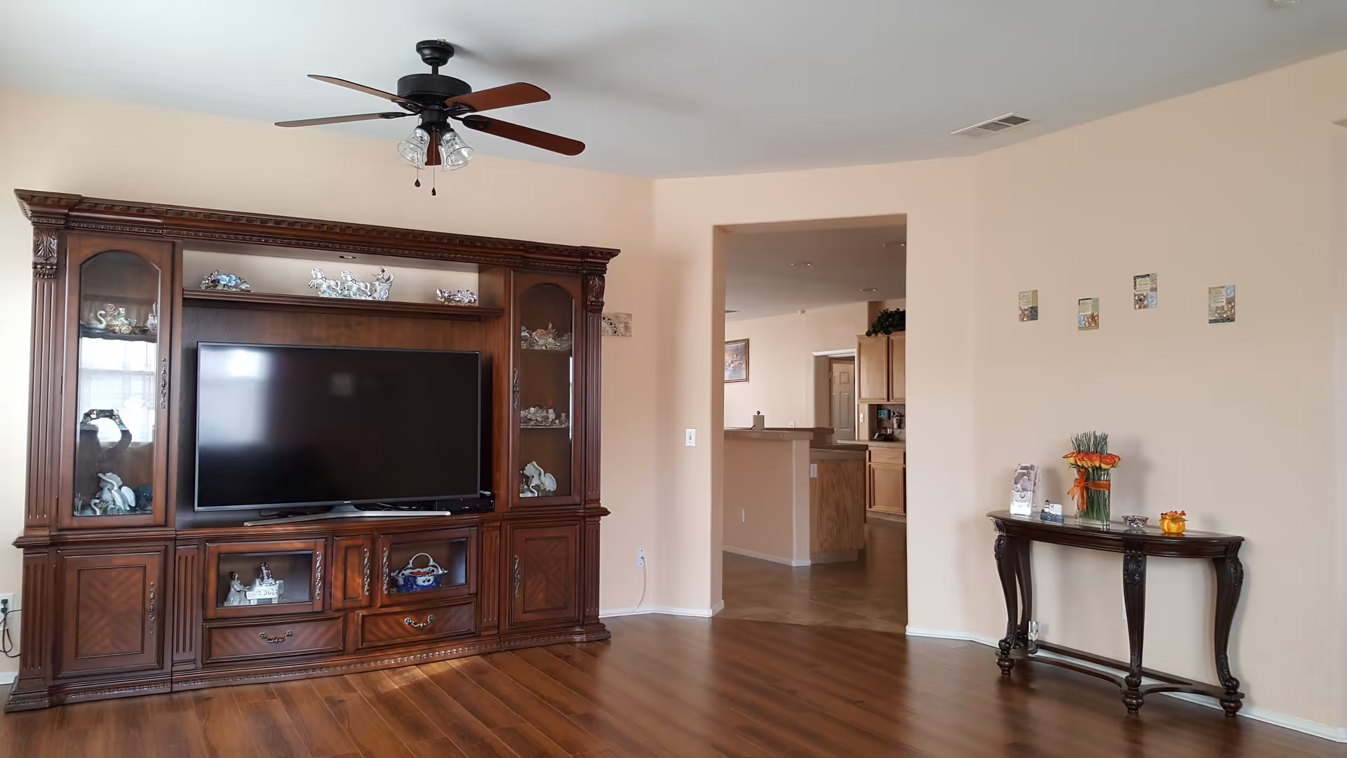 A living room with a large wooden entertainment center housing a flat-screen TV and decorative items. The room has wooden flooring, beige walls, a ceiling fan with lights, and a small wooden console table with flowers and decorative pieces near an open doorway leading to a kitchen area.