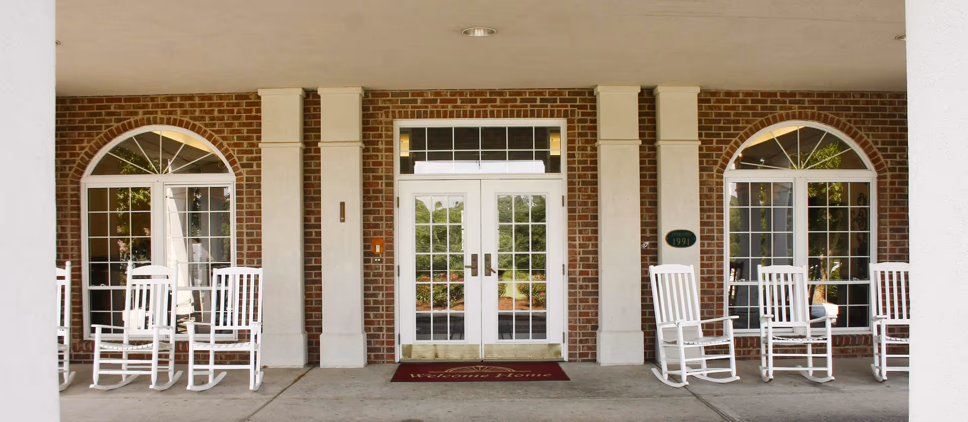 Covered brick entrance with double glass doors flanked by arched windows and white rocking chairs on the porch.