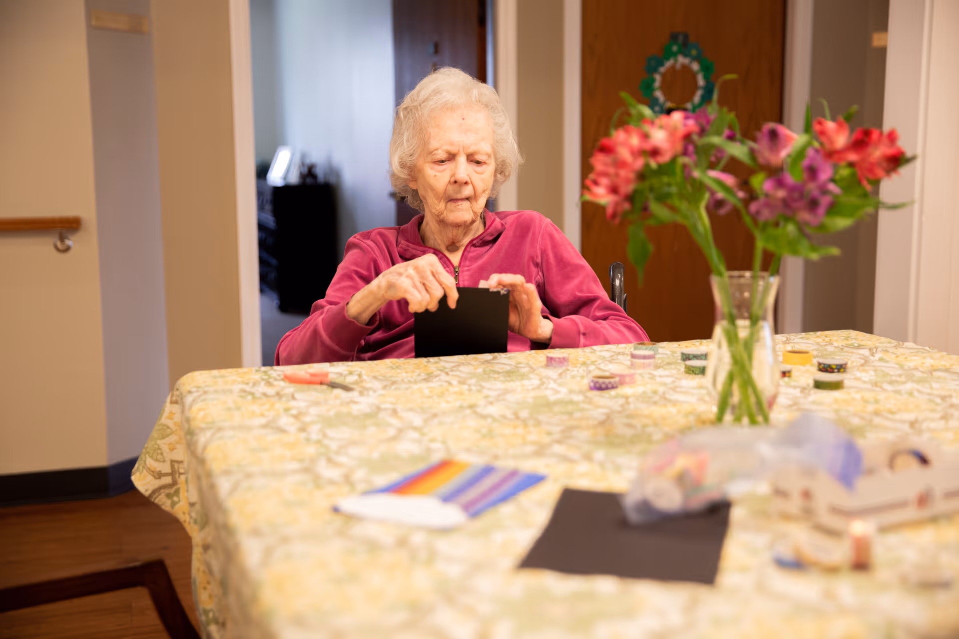 An elderly woman in a magenta jacket sitting at a table covered with a floral tablecloth, engaging in a craft activity with black paper. On the table are various craft supplies including colorful tape rolls, a vase with pink and purple flowers, and other materials. The setting appears to be a cozy indoor room with wooden flooring and a door in the background.