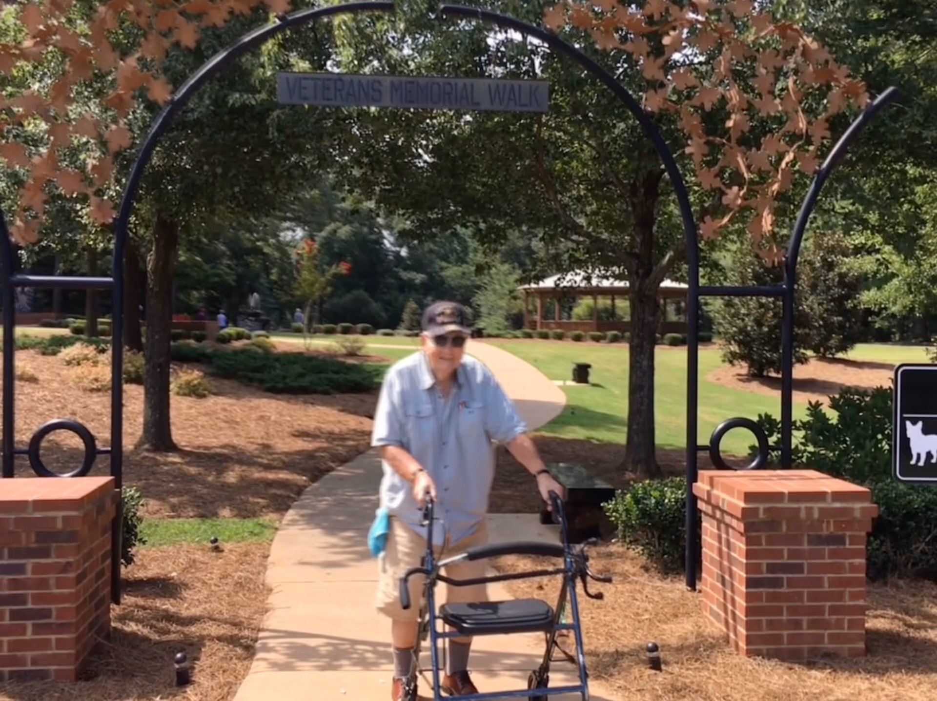 Elderly man with walker walking along paved outdoor path beneath Veterans Memorial Walk arch.