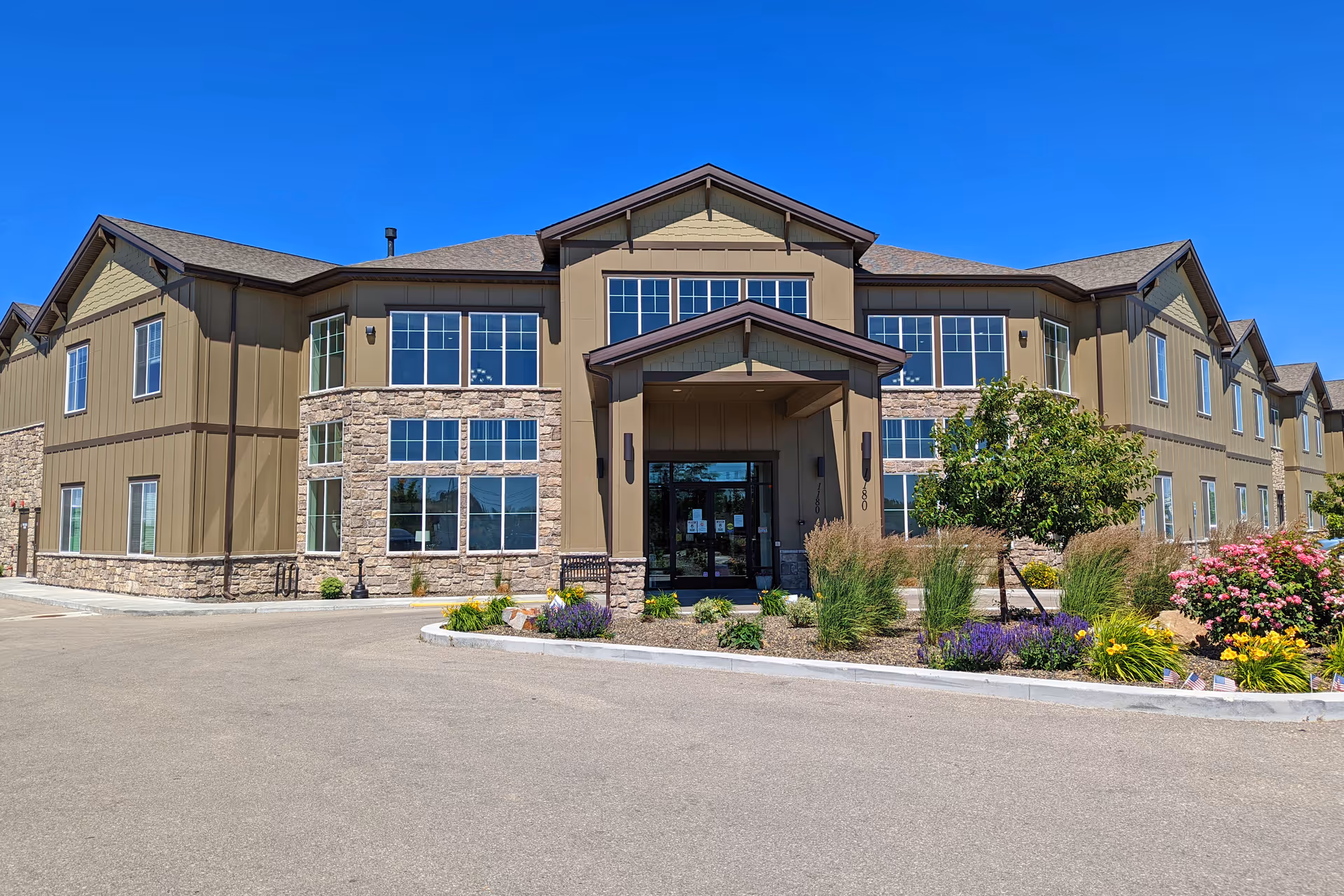 Front exterior of a two-story senior living building with a covered entrance, large windows and landscaped flowerbeds under a clear blue sky.