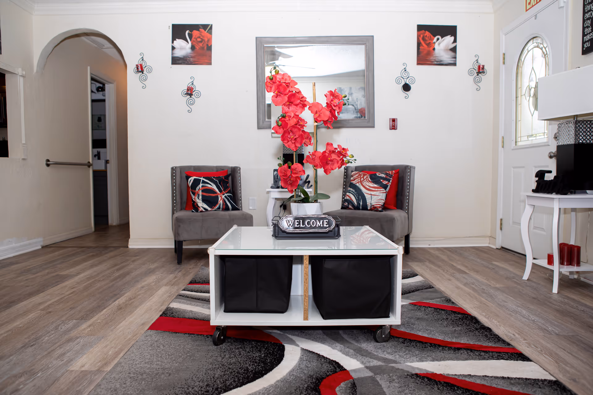 Small reception area with two gray chairs, a white coffee table displaying a 'WELCOME' sign and red flowers, and decorative wall art.
