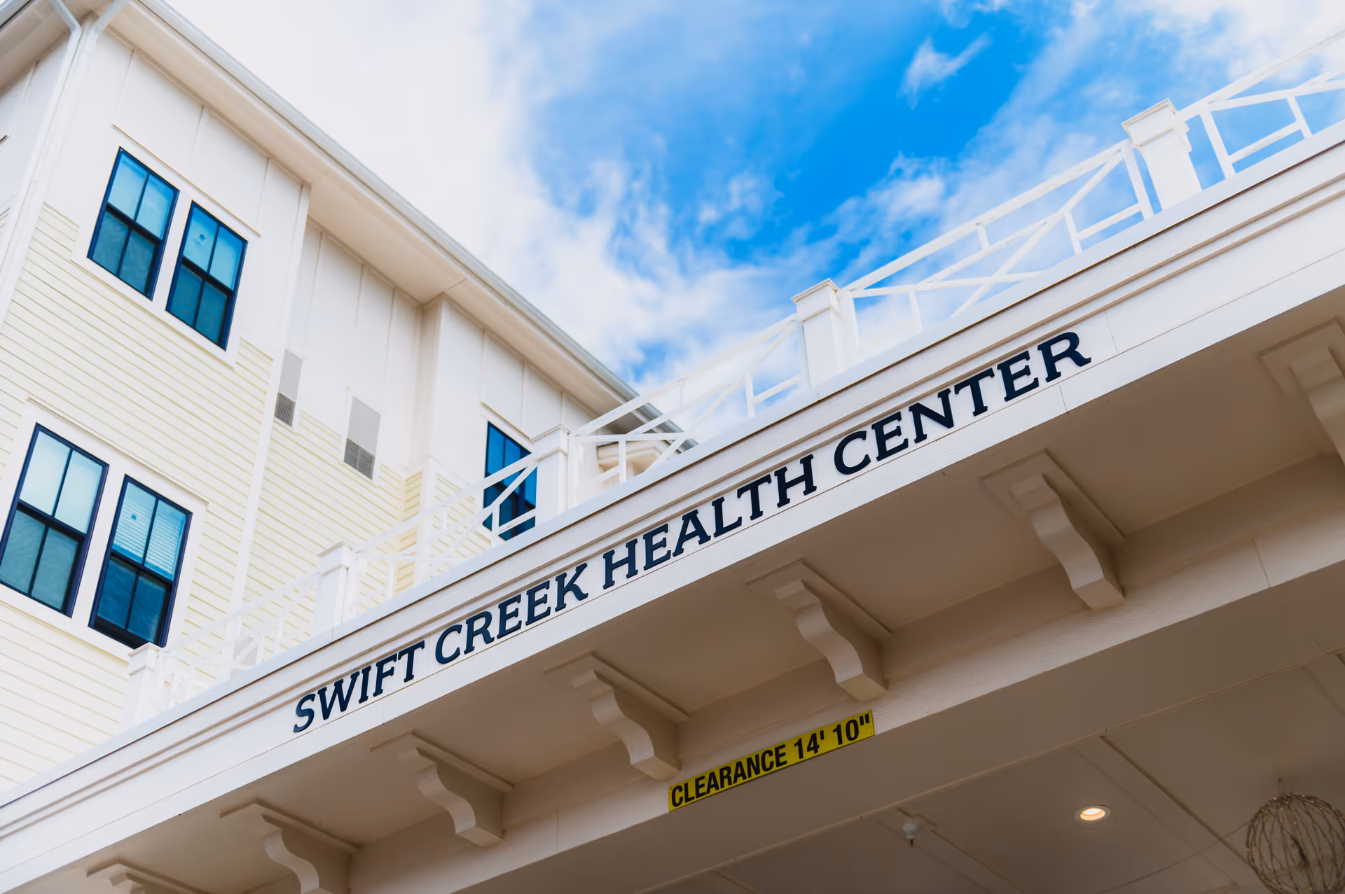 Exterior view of the Swift Creek Health Center building showing part of the facade with windows and a white overhang with the center's name displayed prominently. The sky is blue with some clouds.