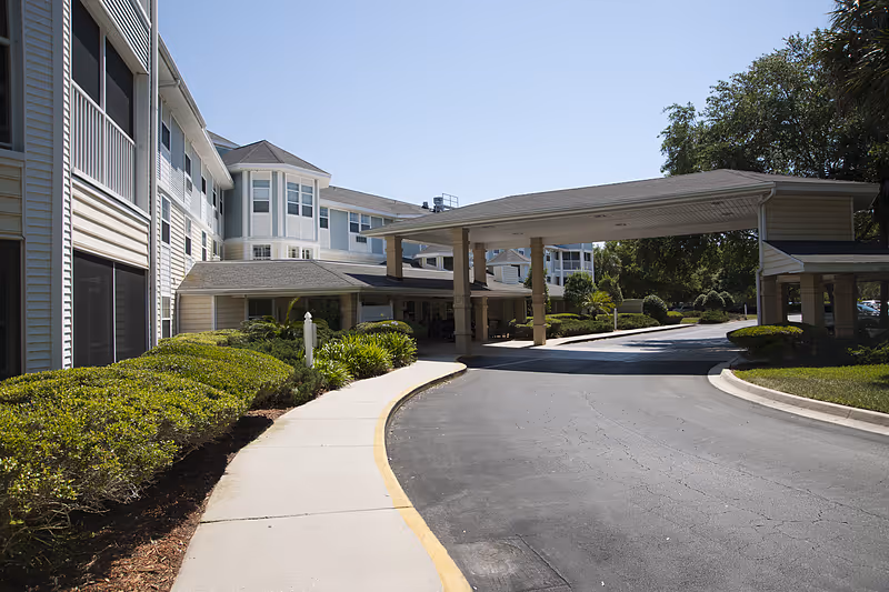 Exterior view of Allegro Senior Living facility in St. Augustine, FL, showing a covered driveway entrance with a curved sidewalk, surrounded by well-maintained bushes and trees under a clear sky.