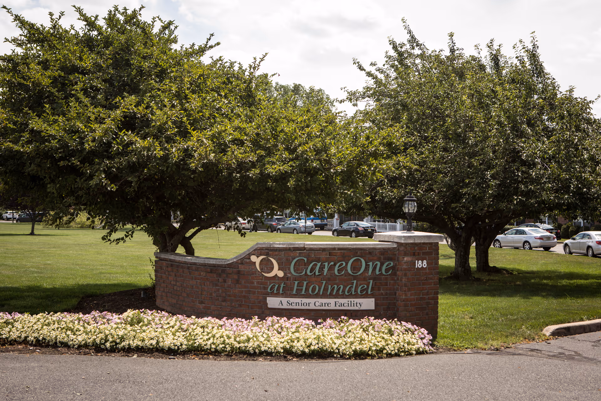 Brick entrance sign reading 'CareOne at Holmdel' surrounded by flowerbeds, trees, and parked cars on the facility grounds.