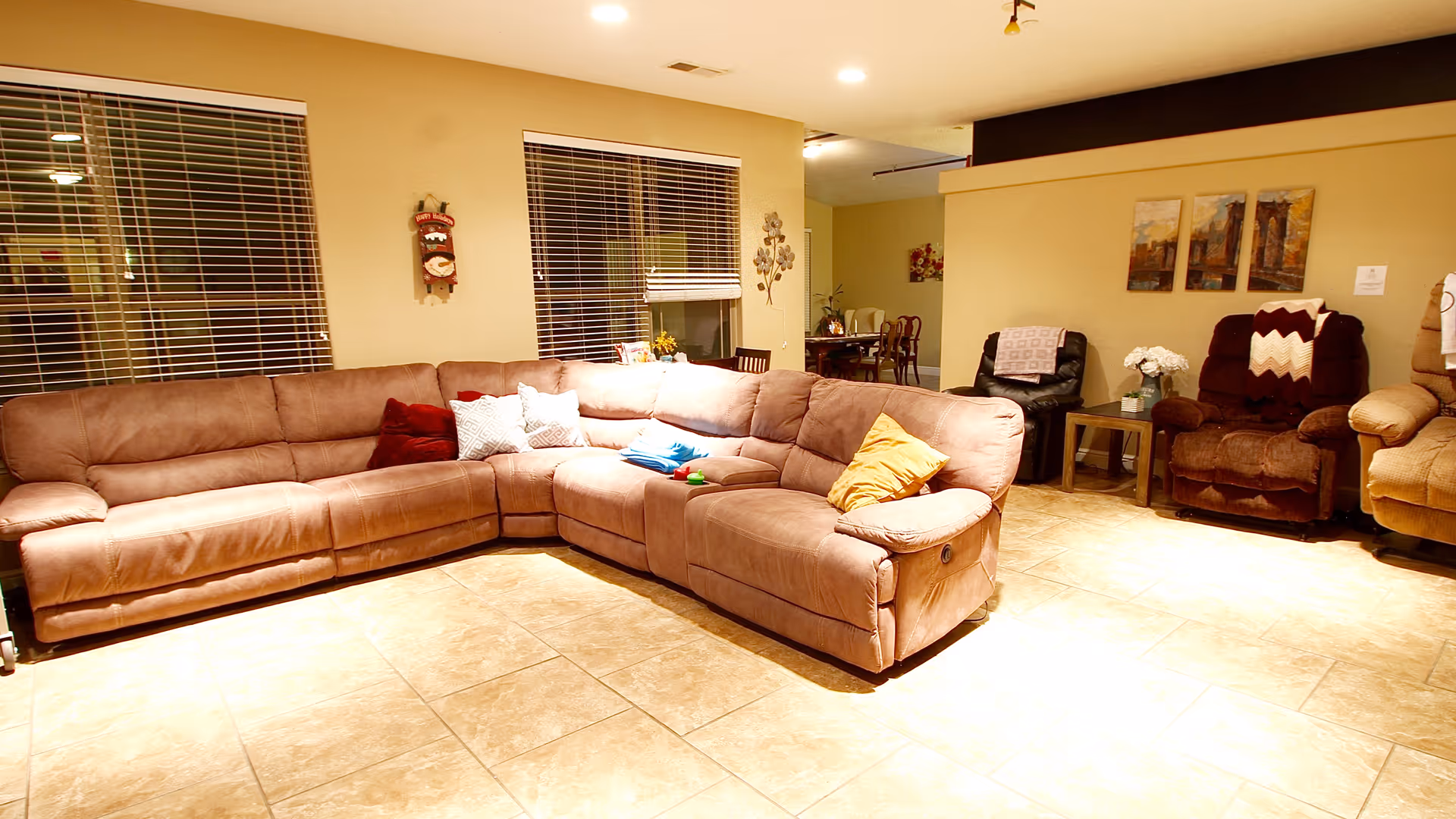 A spacious living room with a large L-shaped brown sectional sofa adorned with various colored pillows. The room has tiled flooring, two windows with blinds, and several armchairs with blankets draped over them. There are decorative wall hangings and paintings, and a dining area is visible in the background.