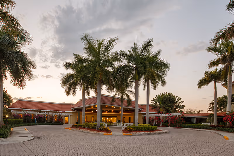 Front entrance of a single-story skilled care building with a circular driveway, tall palm trees, and warm exterior lighting at dusk.