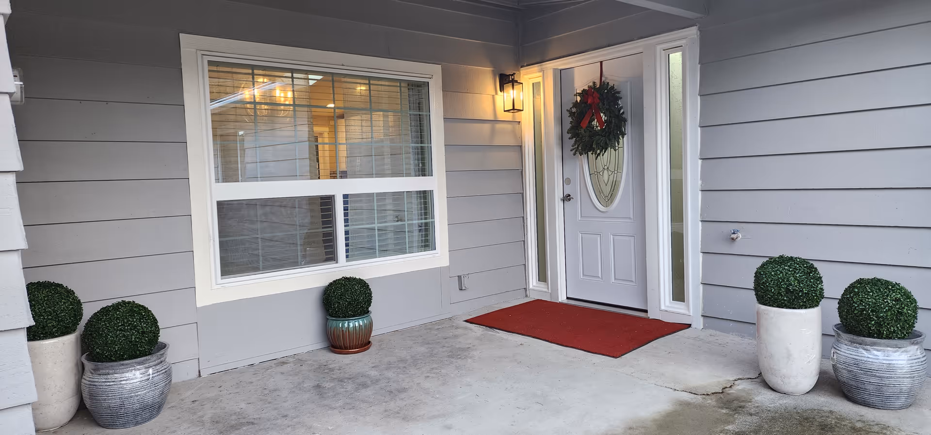 Front entrance of a grey home with wreath, red mat, and potted plants