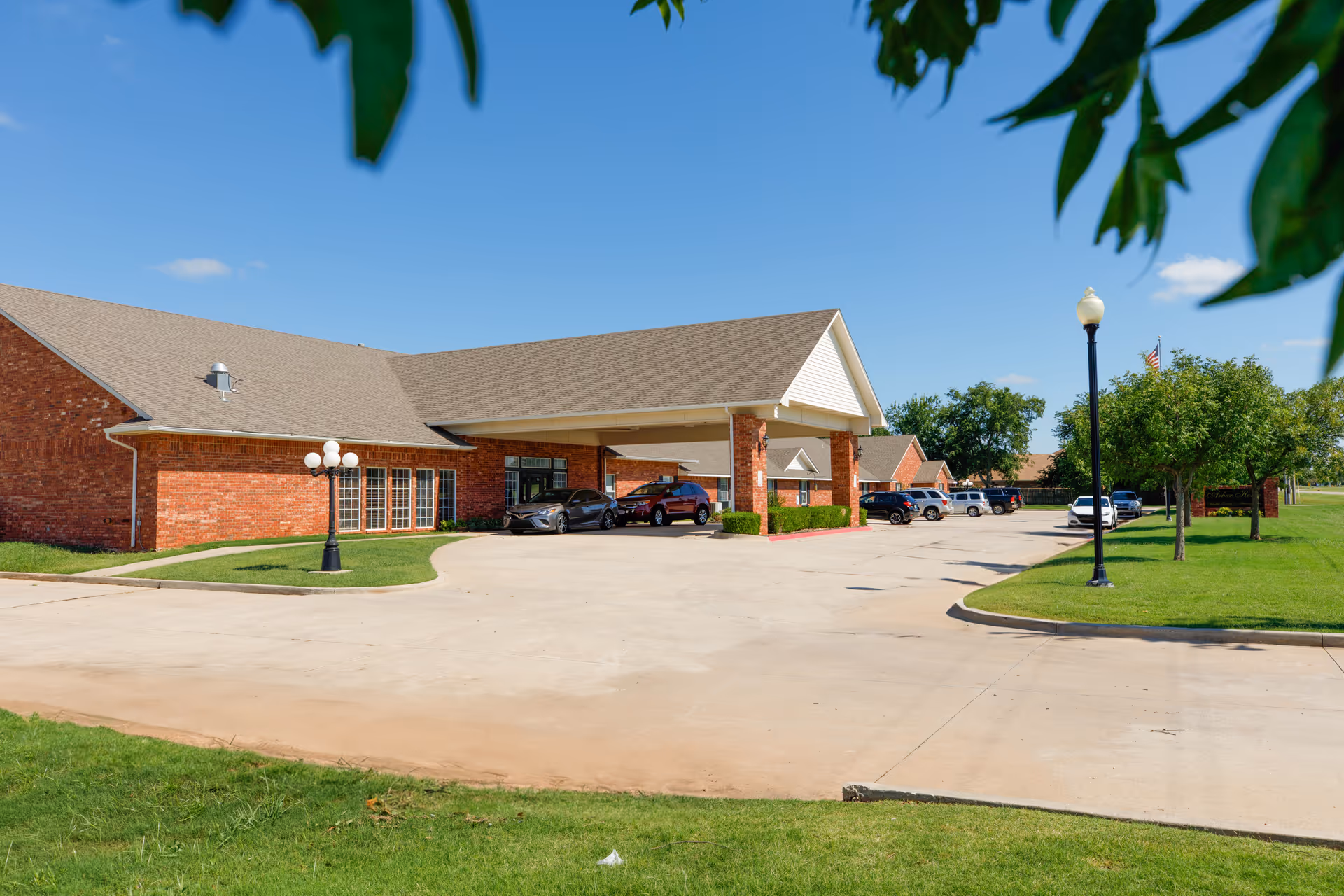 Exterior view of Arbor House Of Mustang, a single-story brick building with a covered entrance and several parked cars in the driveway. The scene includes green grass, trees, and a clear blue sky.