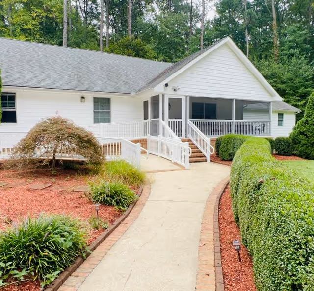 White single-story assisted living entrance with steps, ramp, and manicured landscaping.