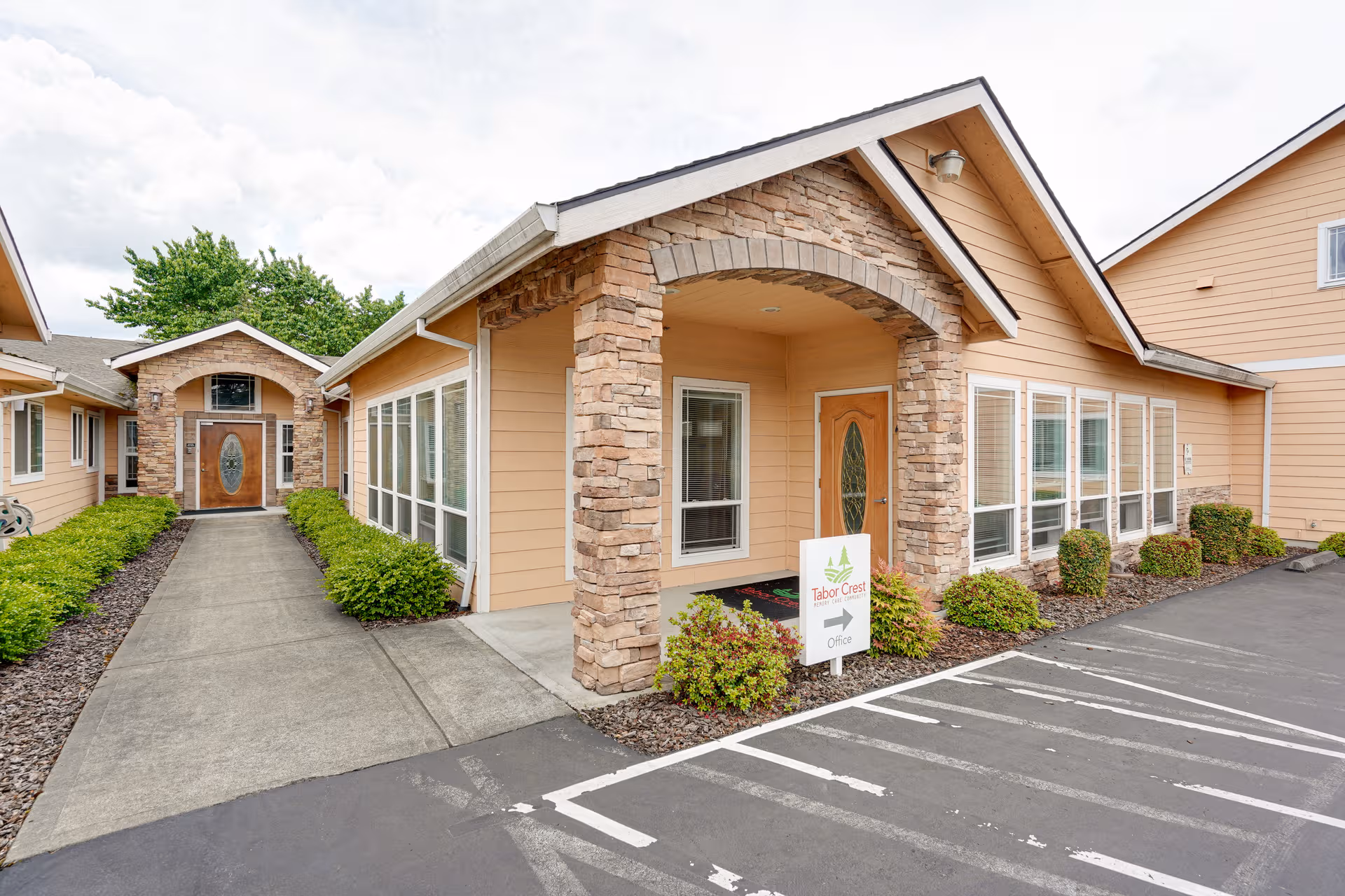 Exterior view of a tan, stone-front senior care facility with entrance and parking.