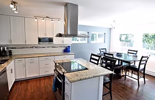 Bright open-plan kitchen with a central island and cooktop, white cabinets, and a dining table by large windows.