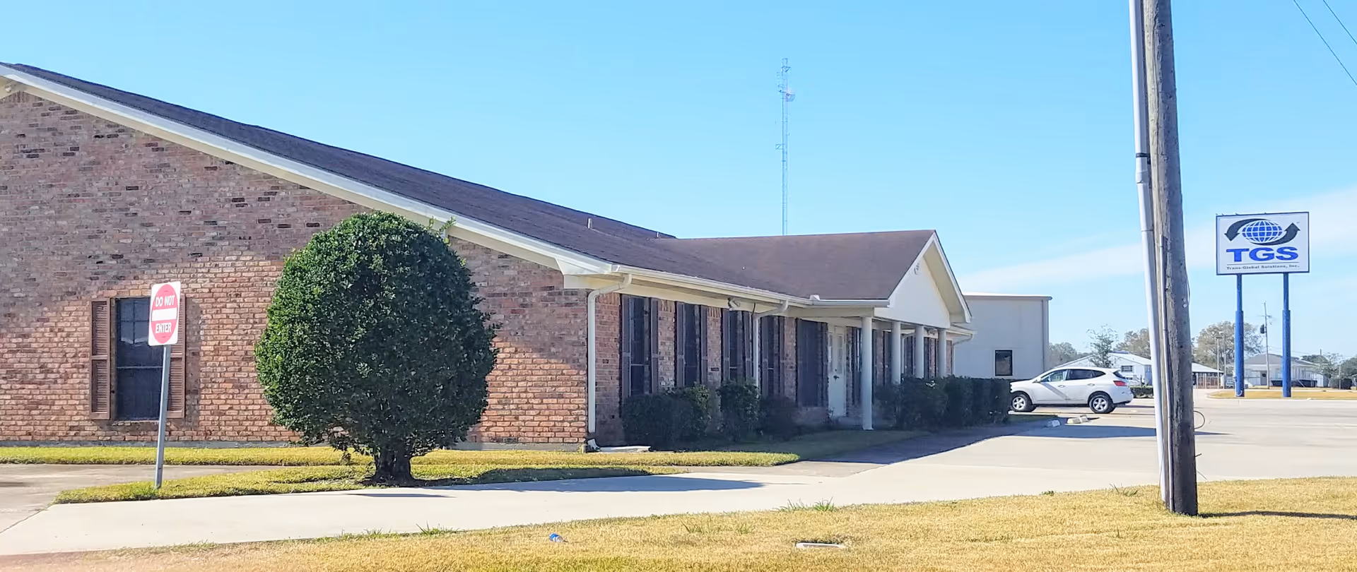 Single-story brick senior housing building with car parking, a trimmed tree, and clear blue sky.