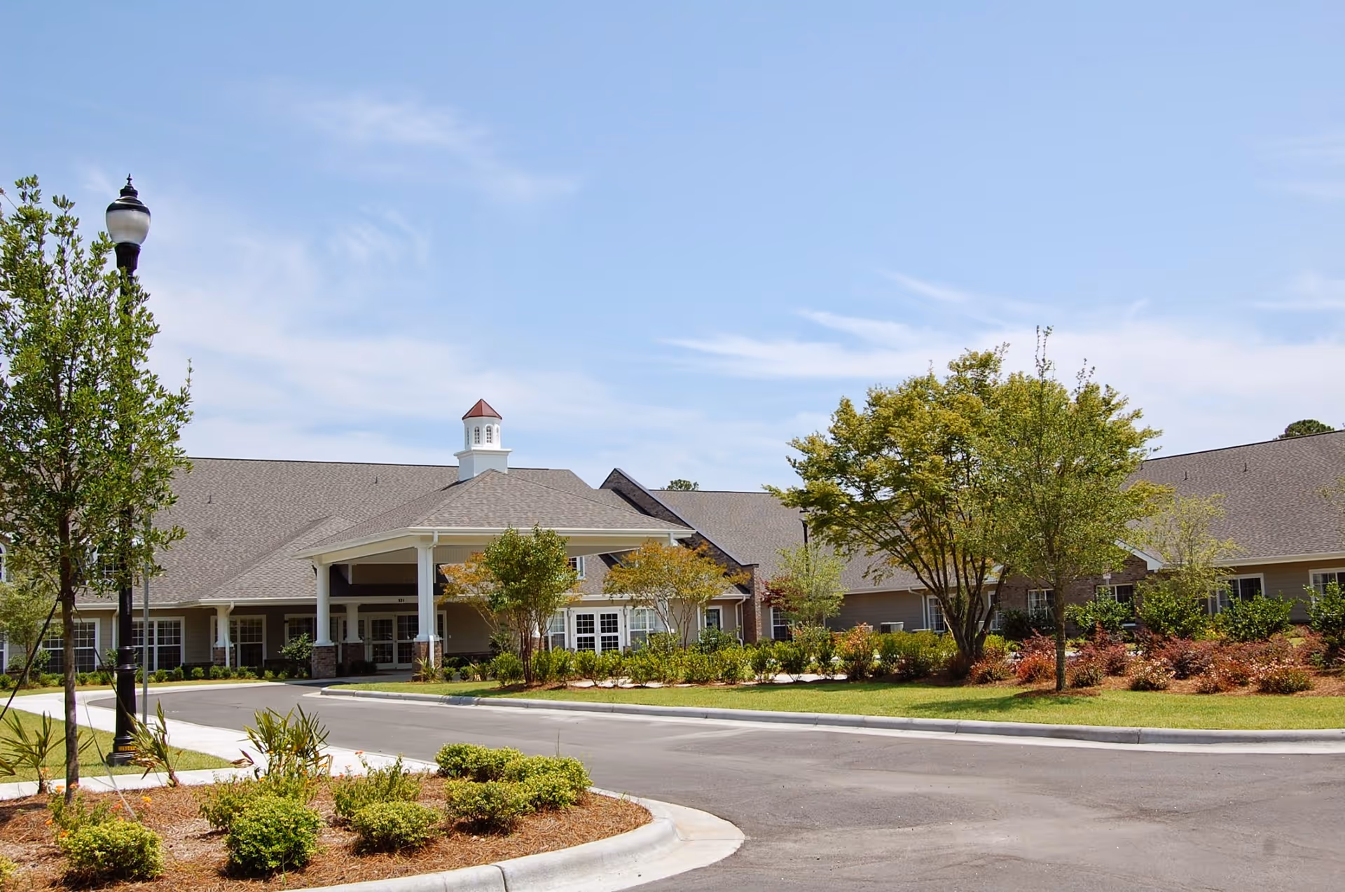Exterior view of a single-story senior living facility building with a covered entrance, surrounded by landscaped greenery and trees under a clear blue sky.