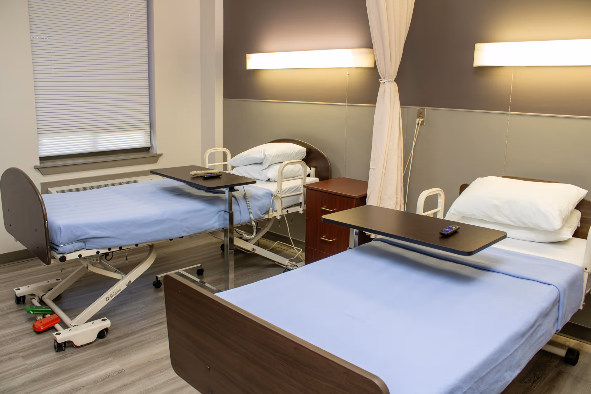 Two hospital beds with tray tables in a well-lit patient room.