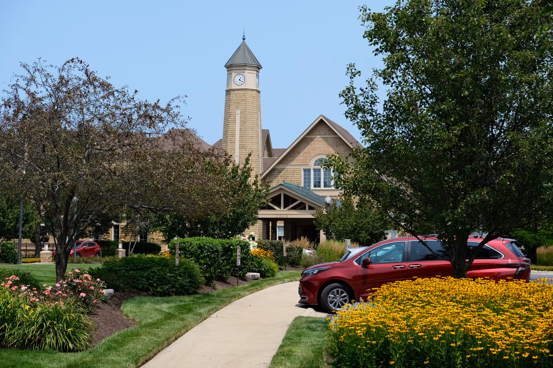 Front exterior of a senior living building with a clock tower, landscaped walkway, flowering beds, trees, and parked red cars.