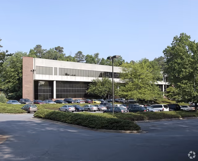 A modern building exterior with a long facade, numerous parked cars, and surrounding trees.