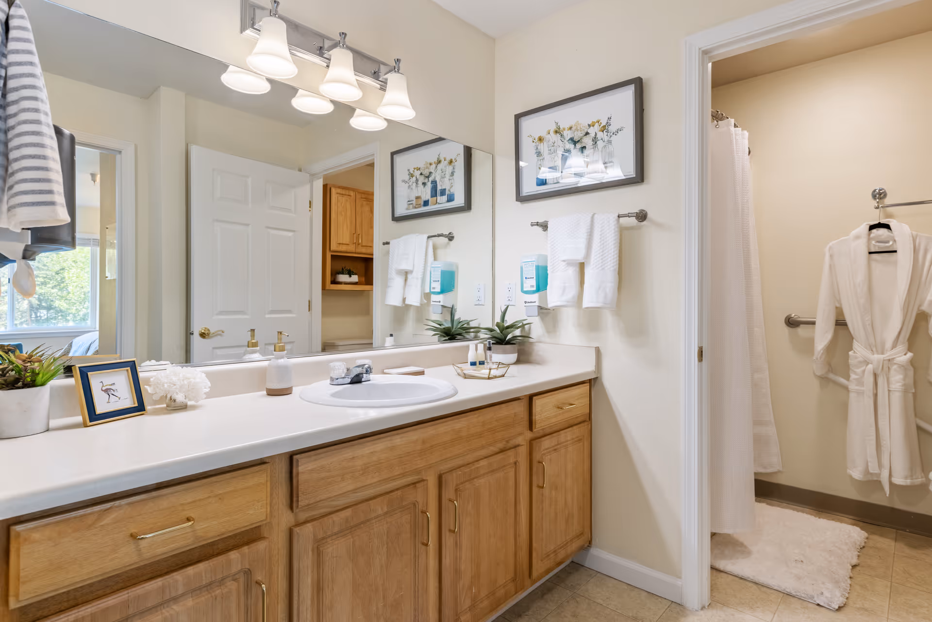 A clean and well-lit bathroom with a large mirror above a wooden vanity with a white countertop and sink. The vanity has gold handles and is decorated with a small framed picture, a potted plant, soap dispensers, and toiletries. Above the mirror are four light fixtures with frosted glass shades. On the wall next to the mirror are two white towels hanging on a towel rack and a wall-mounted soap dispenser. To the right, there is an open doorway leading to a shower area with a white curtain and a white bathrobe hanging on a grab bar. The floor is tiled and there is a white bath mat in front of the shower.