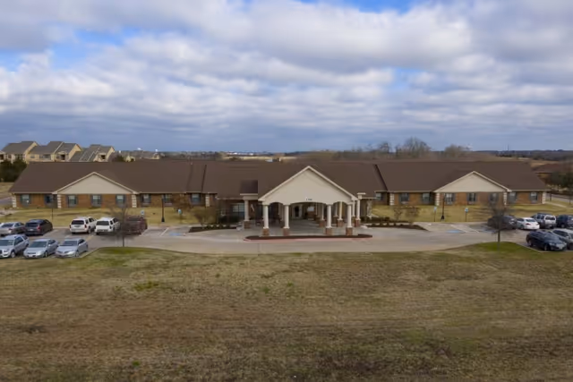 Front exterior view of a single-story healthcare and rehabilitation facility with a covered entrance, multiple windows, and a parking lot with several cars. The building is surrounded by a grassy area under a cloudy sky.
