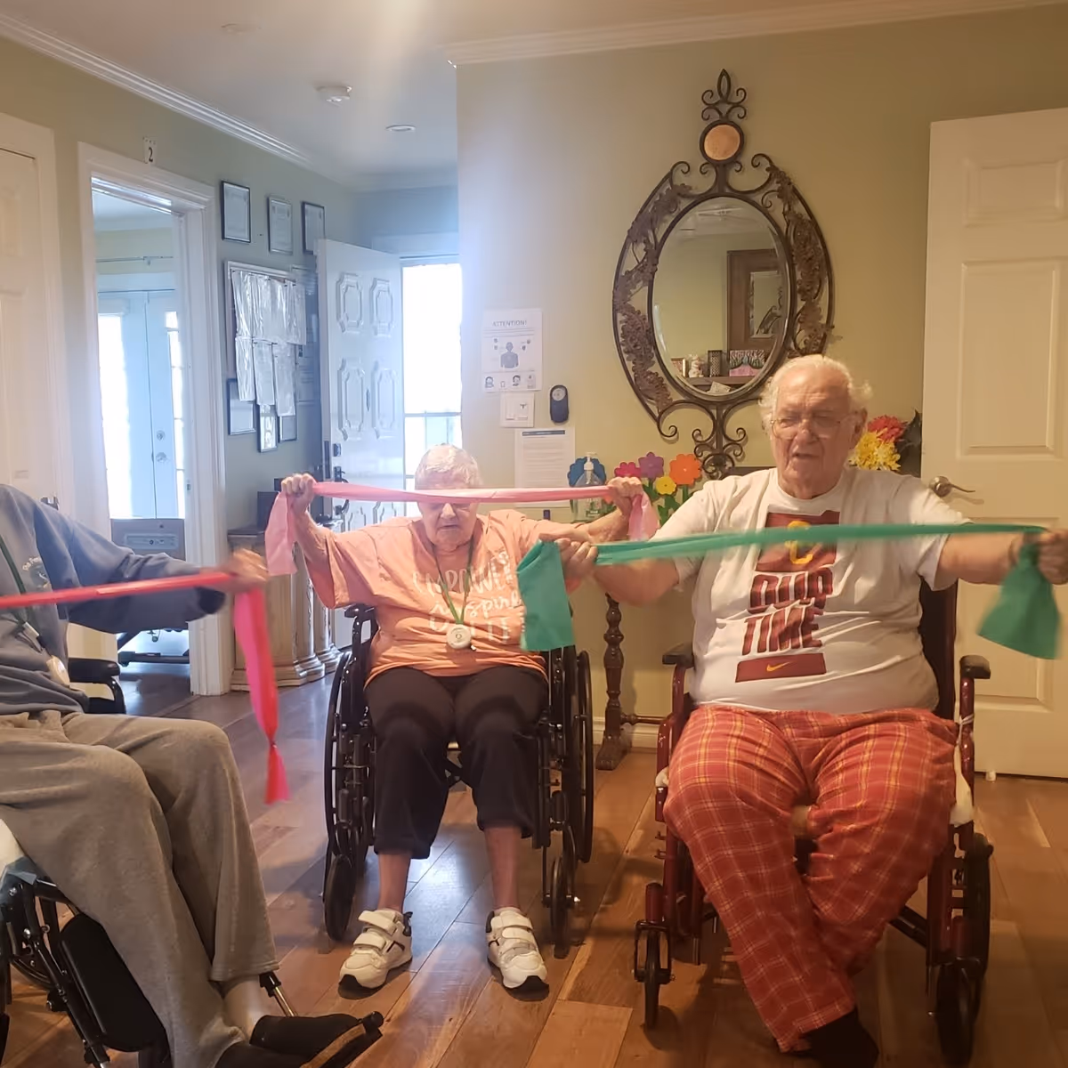 Three elderly individuals sitting in wheelchairs inside a residential care facility, participating in a seated exercise using colorful resistance bands. The room has wooden flooring, a decorative mirror on the wall, and an open door letting in natural light.
