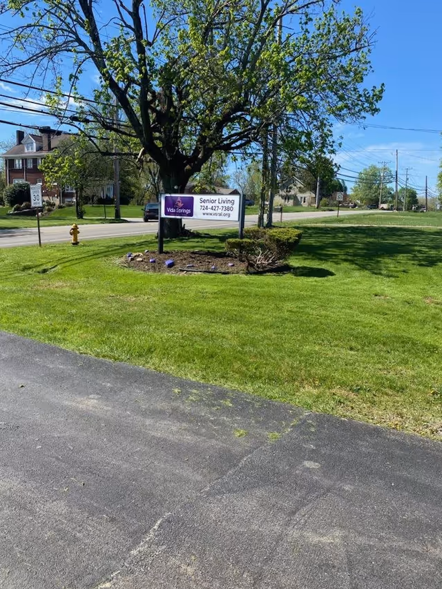 Green lawn and large tree with a Vida Springs Senior Living sign near the roadside on a sunny day.