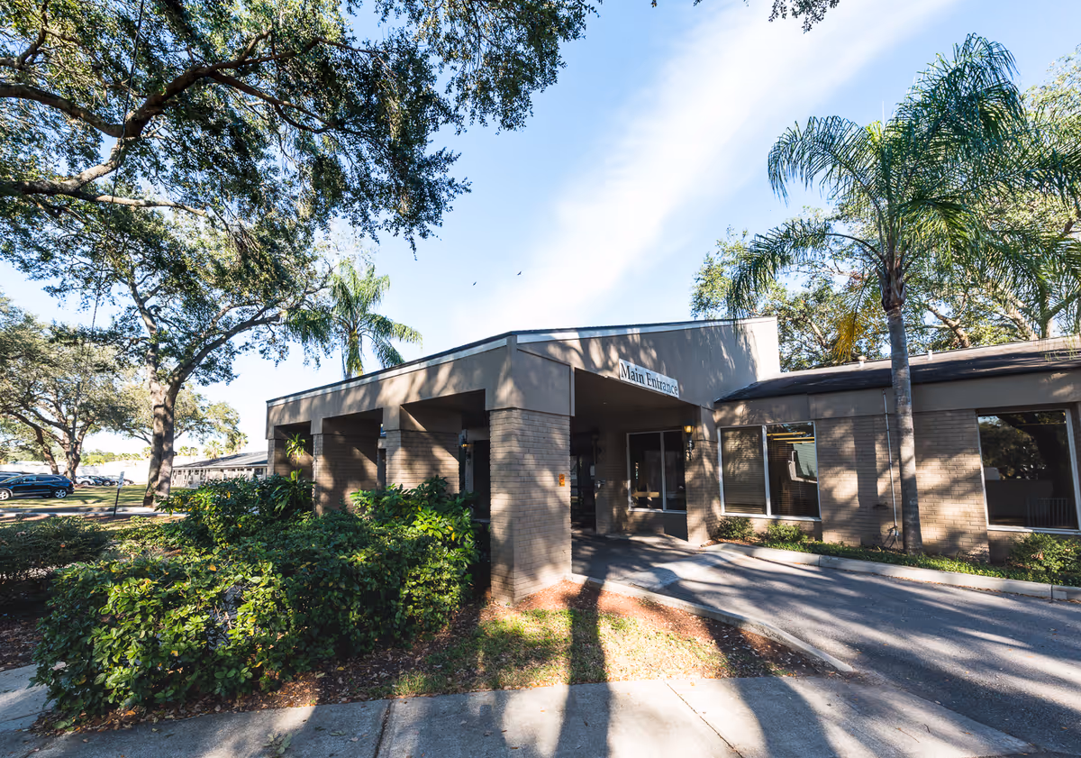 Exterior view of The Bristol at Tampa Rehabilitation & Nursing Center showing the main entrance with a covered driveway, surrounded by palm trees and other greenery under a clear blue sky.