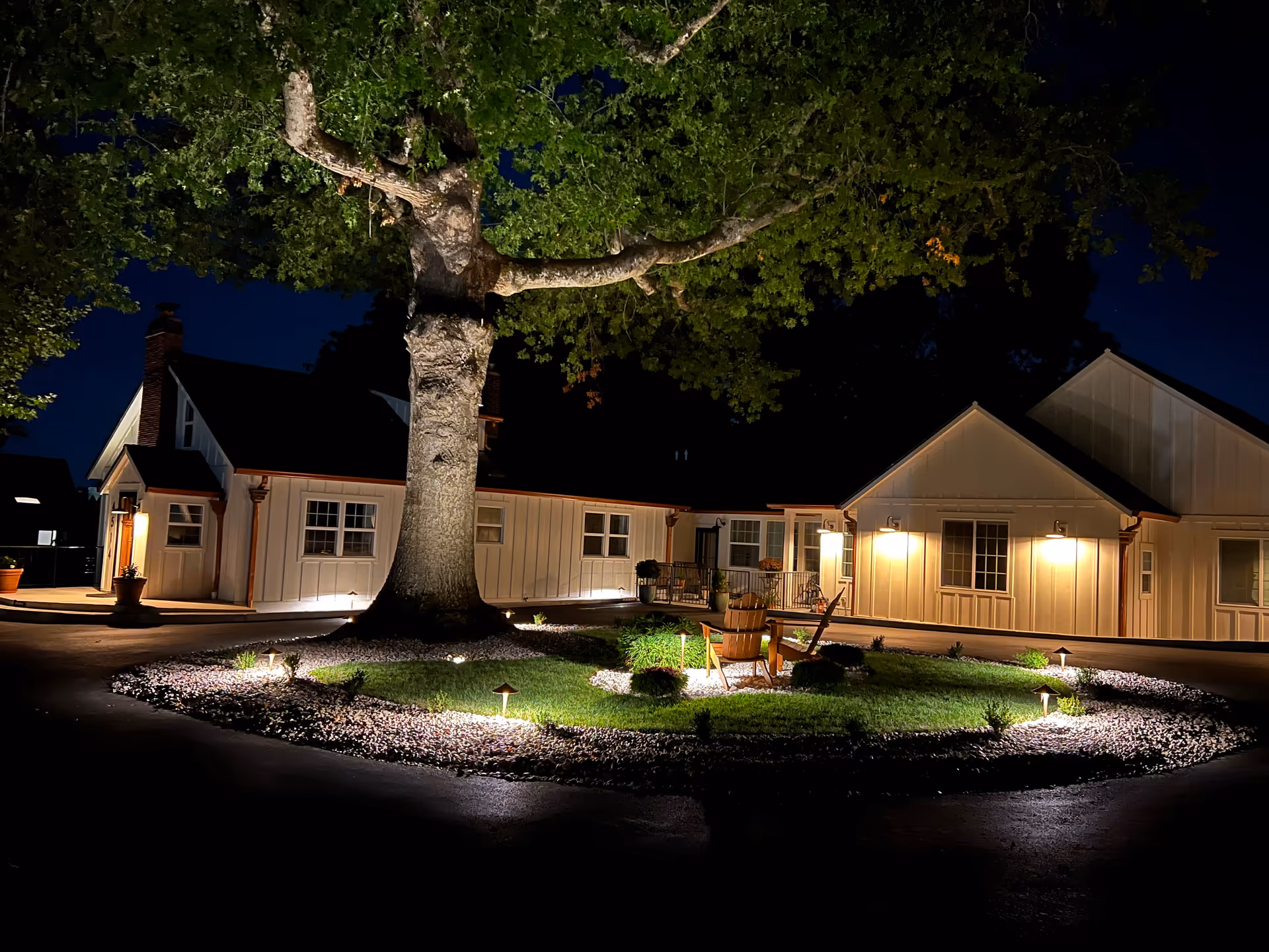 Night exterior of Precious Moments Adult Home Care LLC with a large tree and lit landscaping.