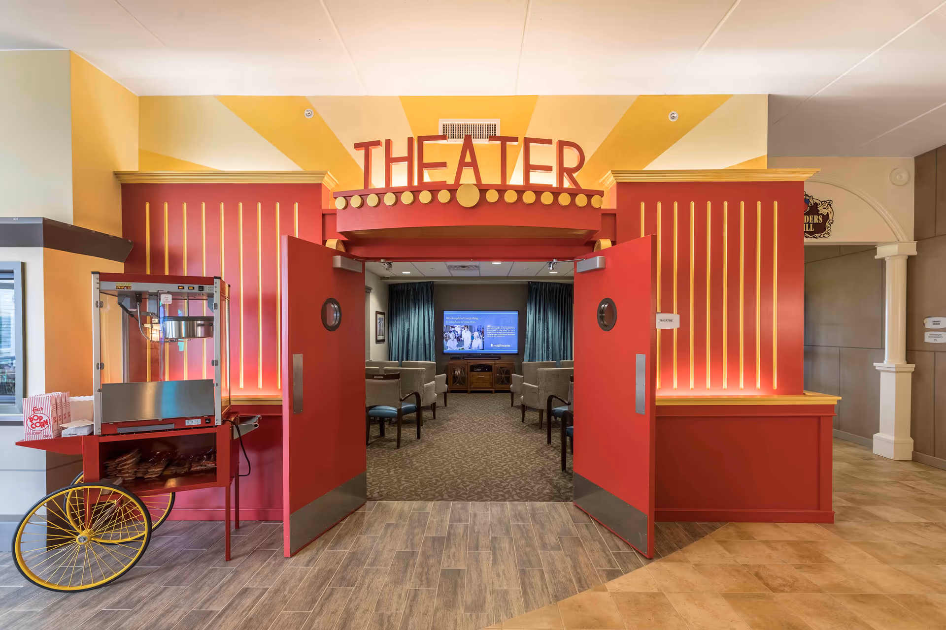 Entrance to a theater room with red double doors open, a red and yellow popcorn machine on the left, and a screen visible inside the room with chairs arranged facing it.
