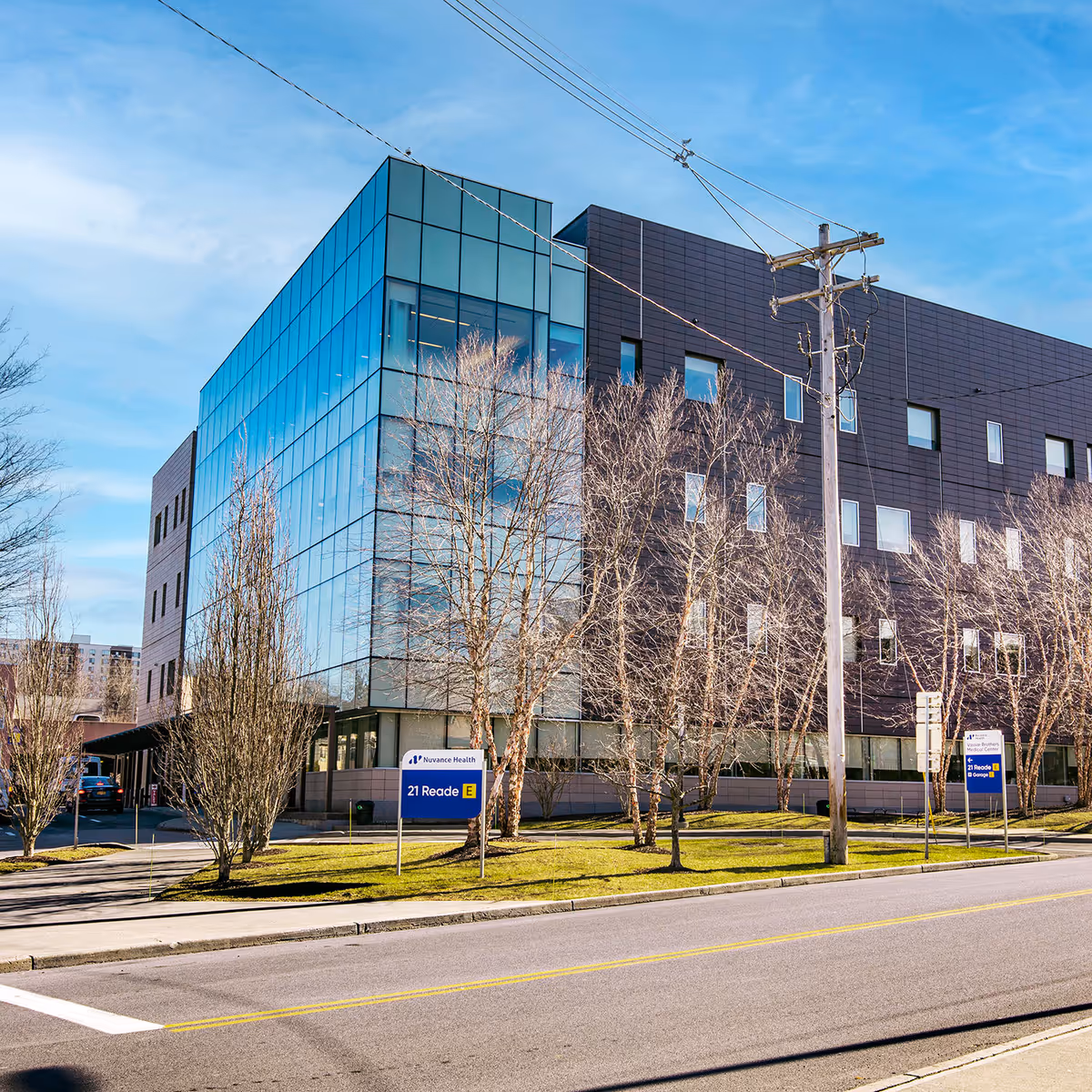 Modern glass-front medical building with blue windows, signage, leafless trees, and street view.