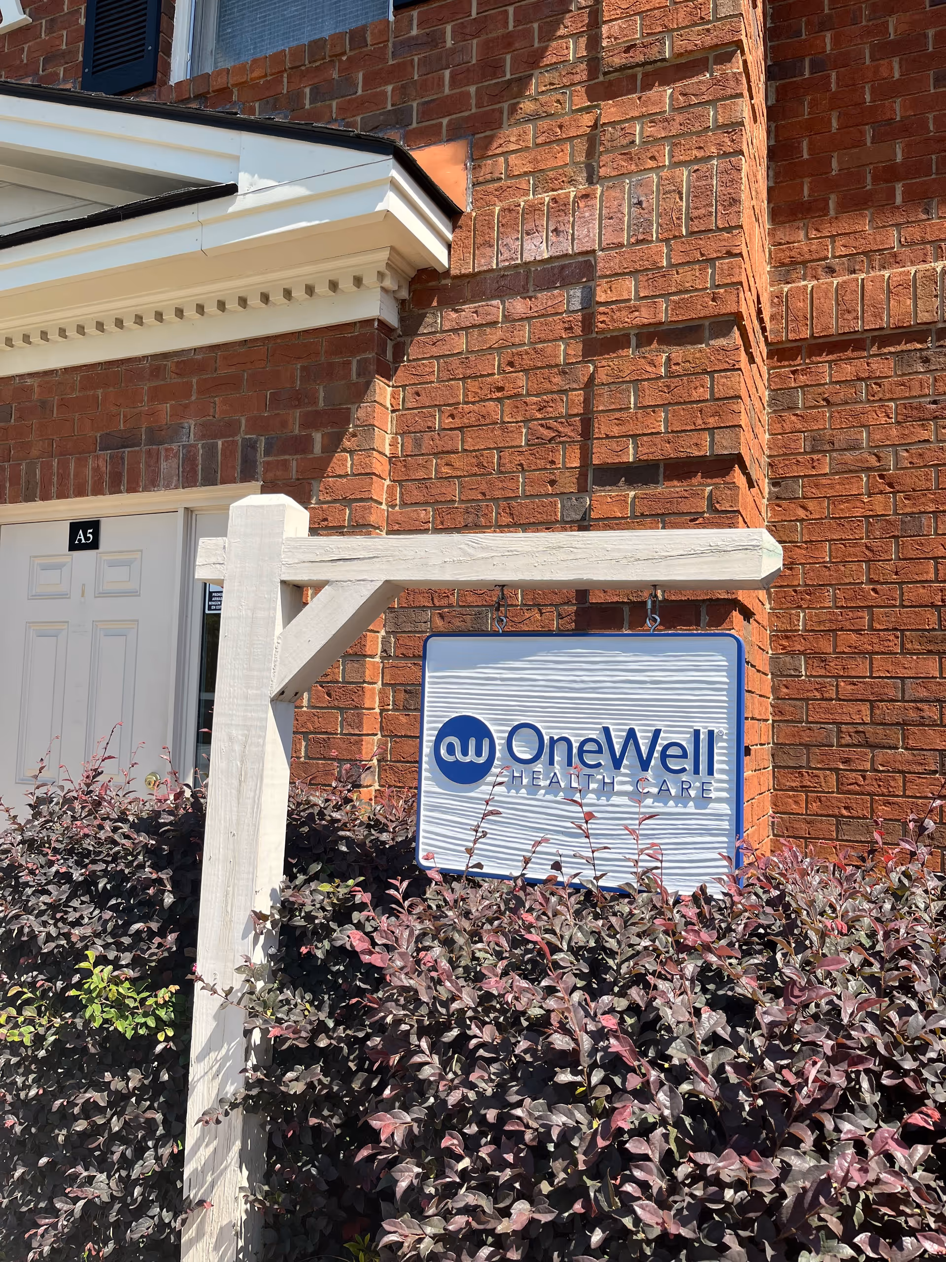 Exterior brick building with a OneWell Health Care sign near shrubs.