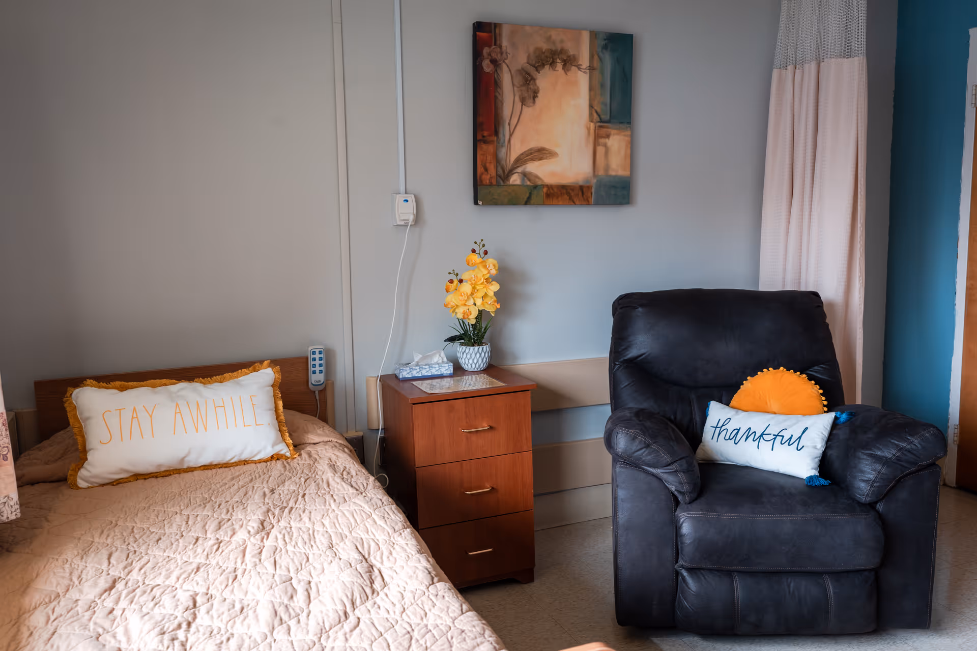 A cozy senior care room with a bed, bedside table topped with flowers and tissues, and a recliner chair with decorative pillows.