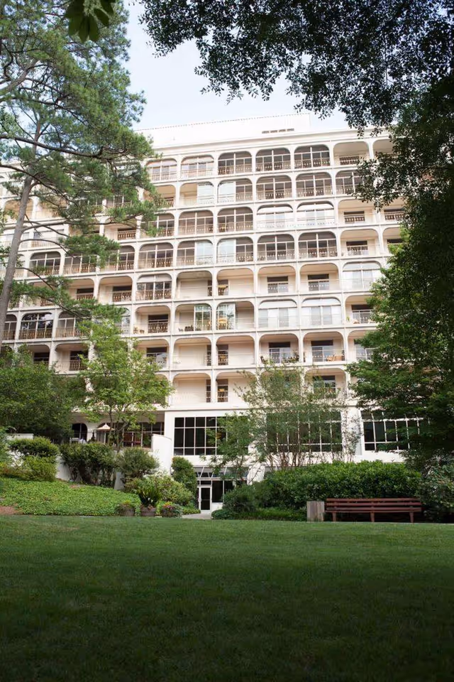 Exterior view of a multi-story senior living facility named Canterbury Court, surrounded by trees and greenery with a well-maintained lawn and a wooden bench in the foreground.