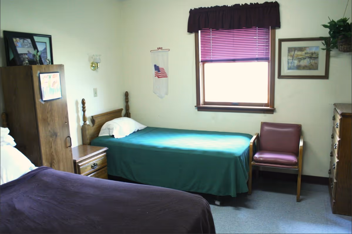 A simple bedroom with two single beds, one with a green bedspread and the other with a dark purple bedspread. There is a wooden nightstand between the beds, a wooden dresser on the right, a wooden cabinet on the left with framed photos on top, a maroon chair near the window, and a window with purple blinds and a valance. The walls are light-colored with a small American flag decoration and a framed picture.