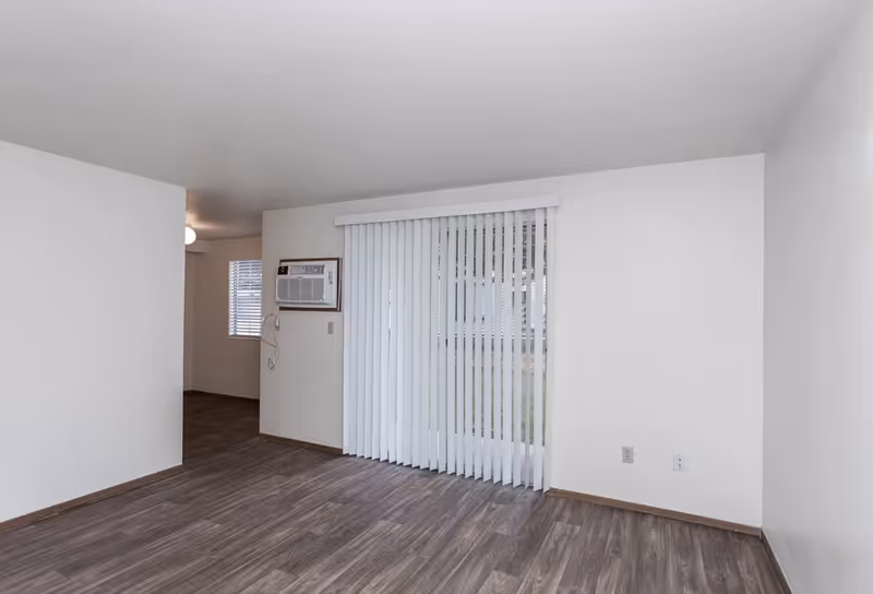 Empty living room with white walls, vertical blinds, and wood-like flooring.