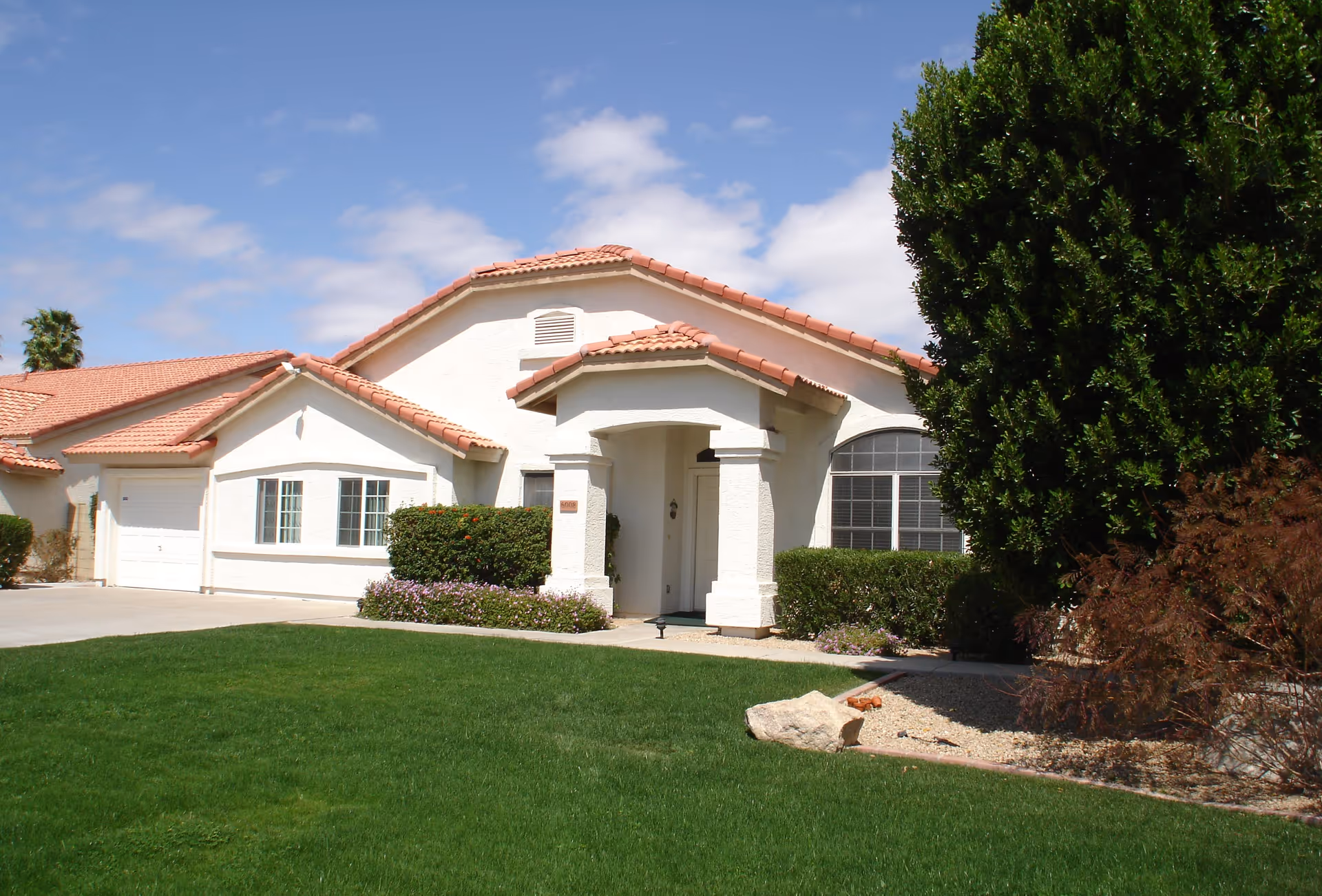 Single-story white stucco house with a red tile roof, green front lawn and driveway under a blue sky.