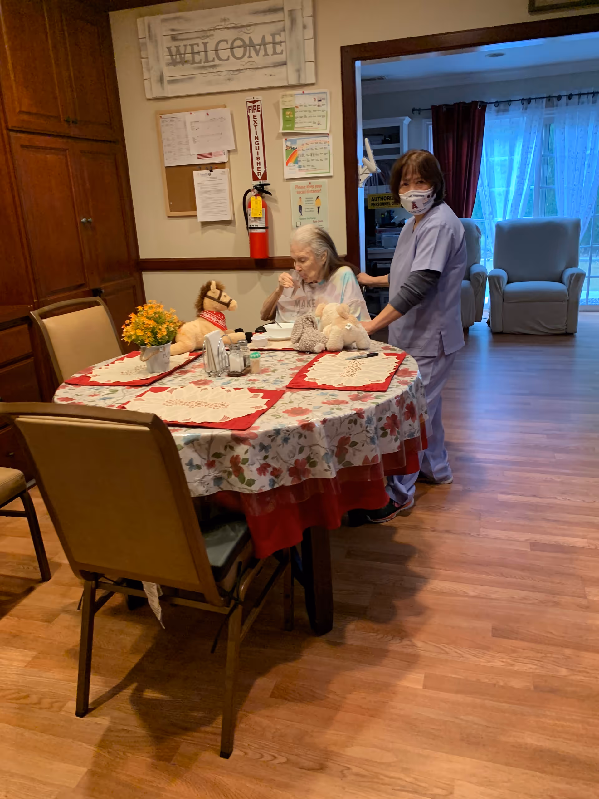 An elderly woman sitting at a round dining table with a floral tablecloth and red placemats, accompanied by a caregiver wearing scrubs and a face mask. The table has stuffed animals and a small flower pot on it. Behind them is a wall with a 'WELCOME' sign, a fire extinguisher, and some notices. In the background, there are two armchairs near a window with curtains.