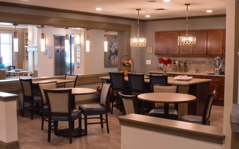A communal dining area with round tables and chairs, a kitchen island, pendant lights, and a seating area visible through a wide interior opening.