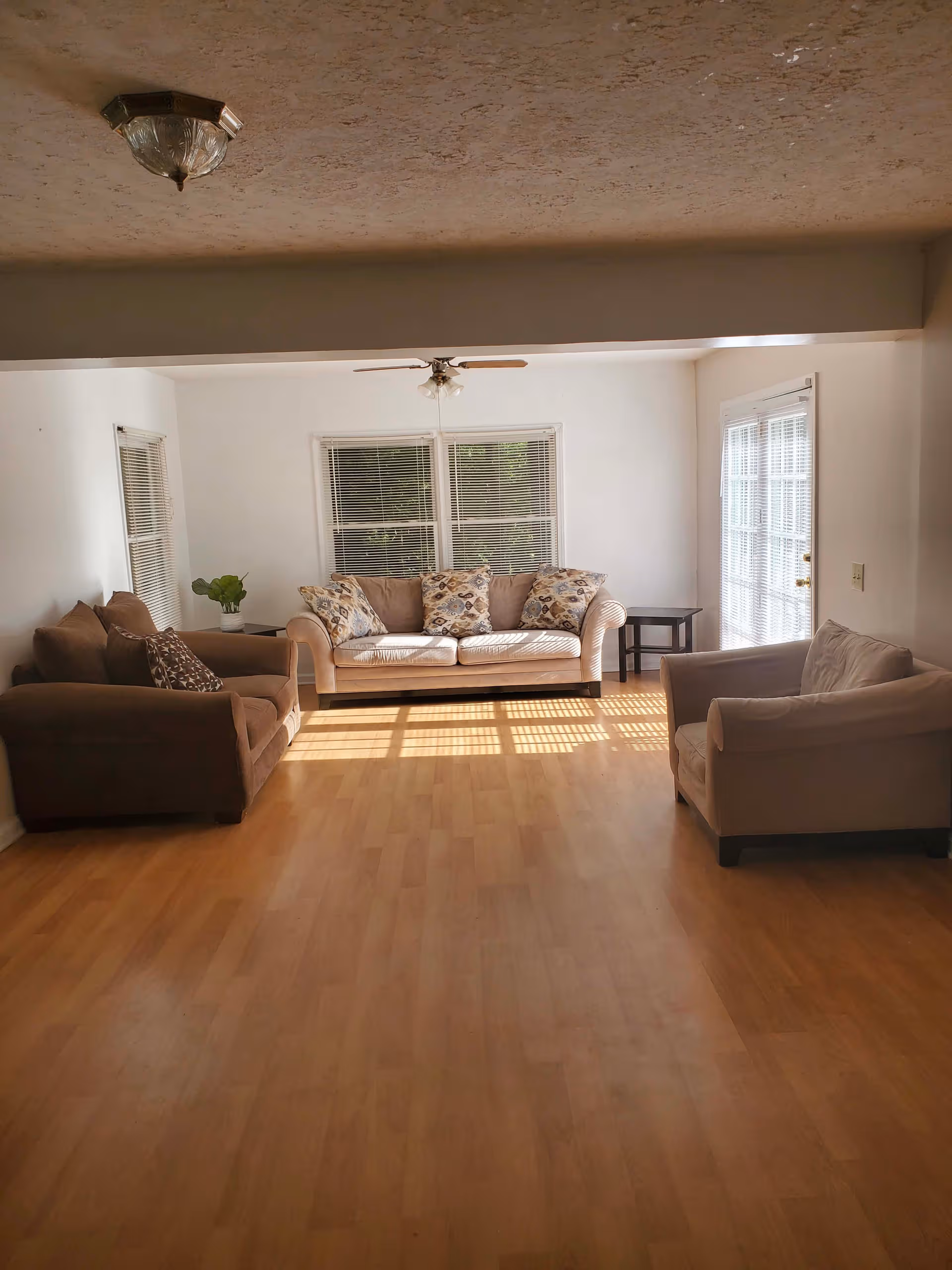Sunlit living room with beige couches, cushions, windows, and ceiling fans.