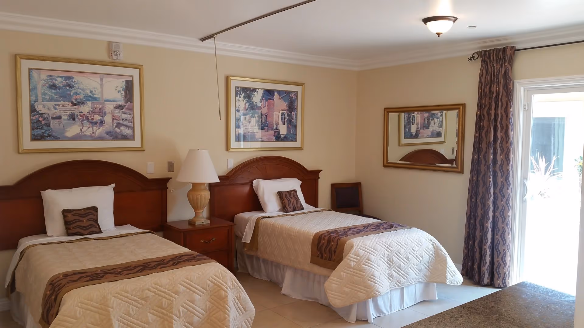 A bedroom with two single beds, each with a wooden headboard, beige bedspreads, and decorative pillows. Between the beds is a wooden nightstand with a lamp. The walls are light-colored and decorated with framed paintings. There is a large mirror on the right wall and a window with patterned curtains letting in natural light.