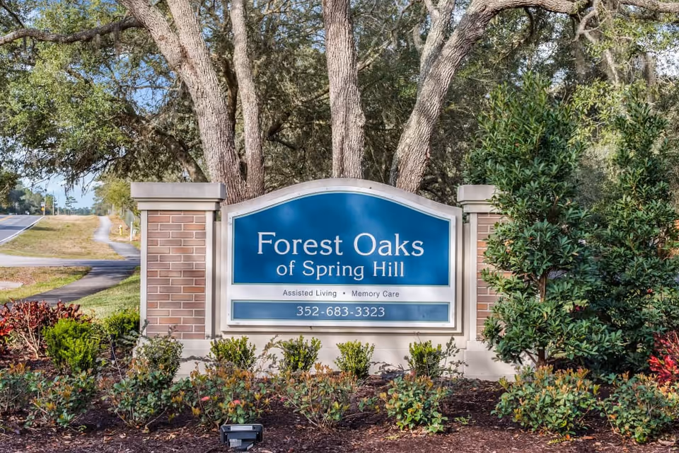 Entrance sign for Forest Oaks of Spring Hill assisted living and memory care facility, surrounded by trees and landscaping with a road visible in the background.