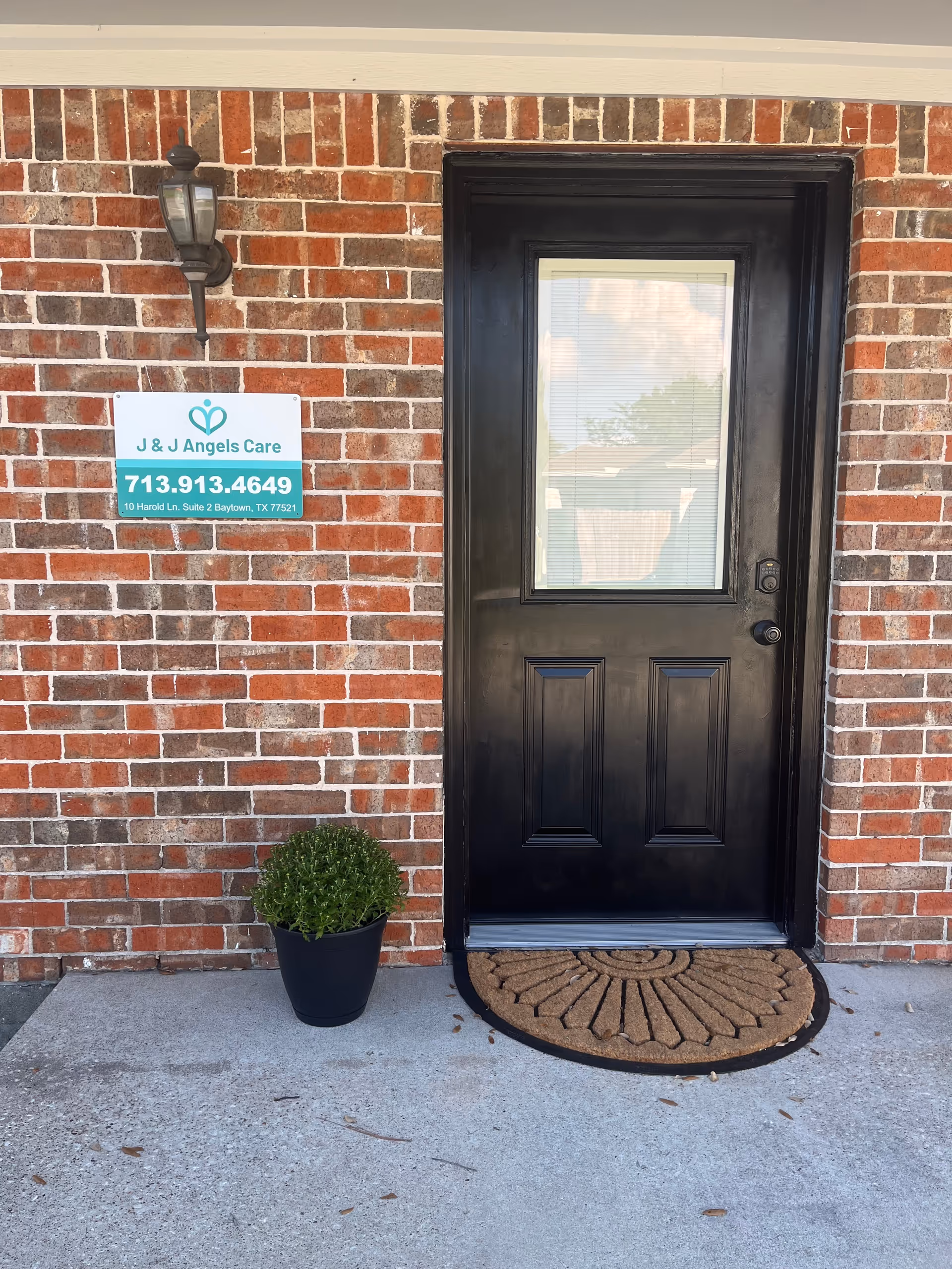 Brick building exterior with black front door, mat, plant, and J & J Angels Care sign.