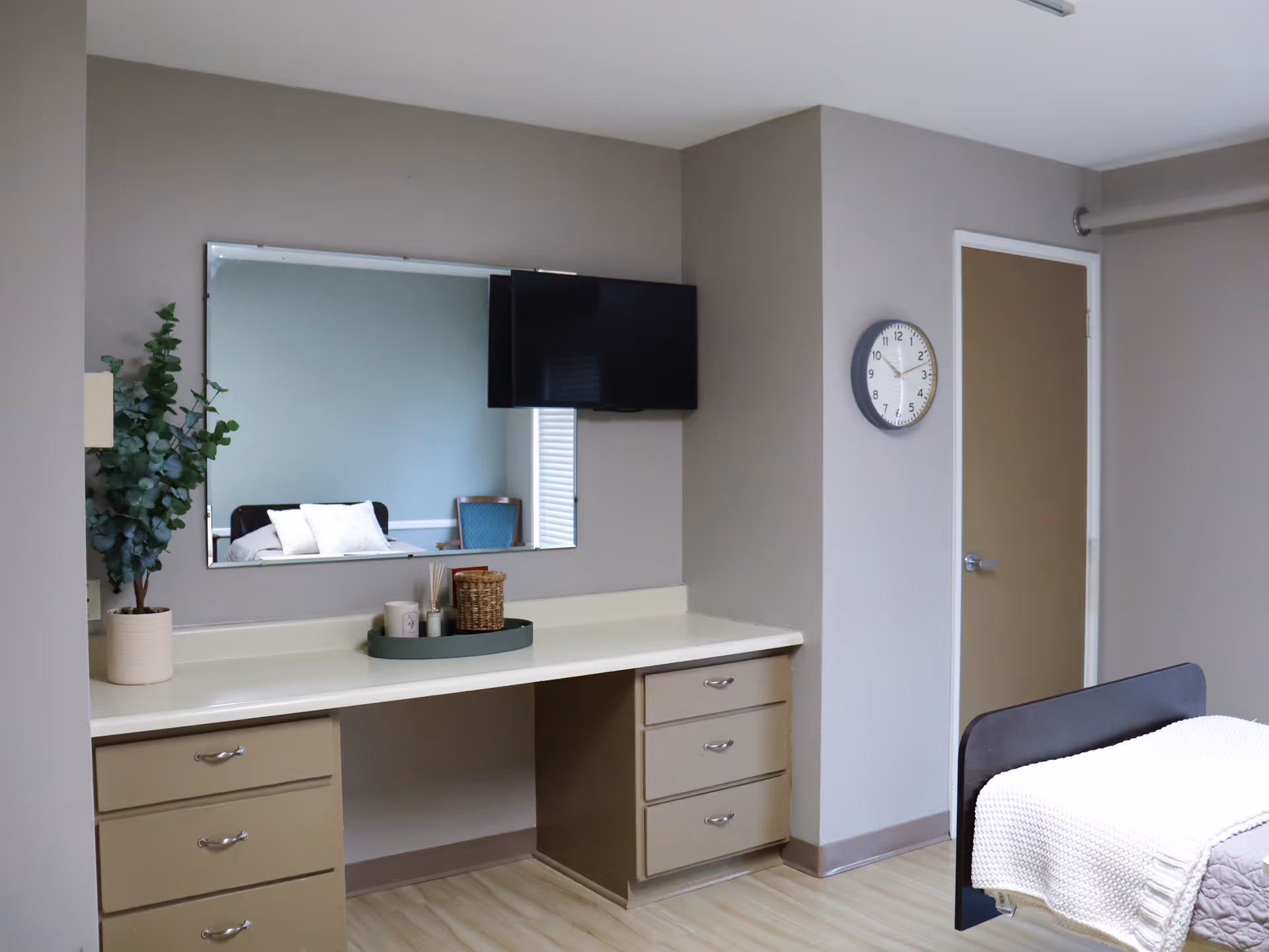 Furnished bedroom with a built-in desk and drawers, wall-mounted TV and mirror, clock on the wall, and a bed with a white blanket.