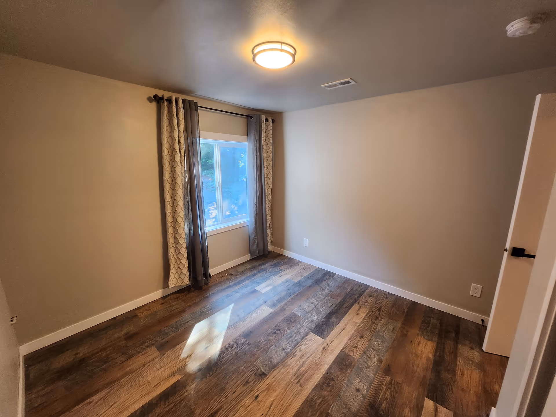 Empty room with wooden flooring, beige walls, a window with patterned curtains, and a ceiling light fixture.