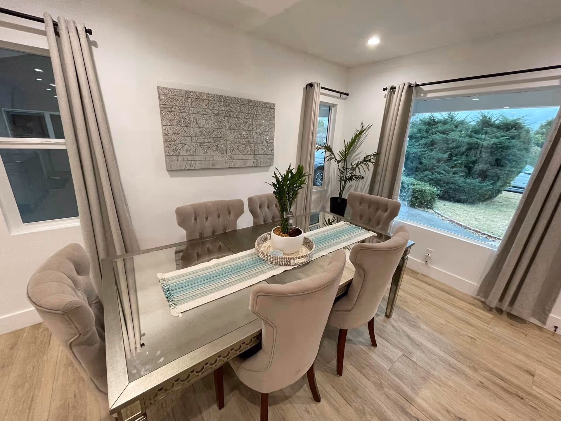 Glass dining table with beige tufted chairs, plants, and windows in a bright dining room.