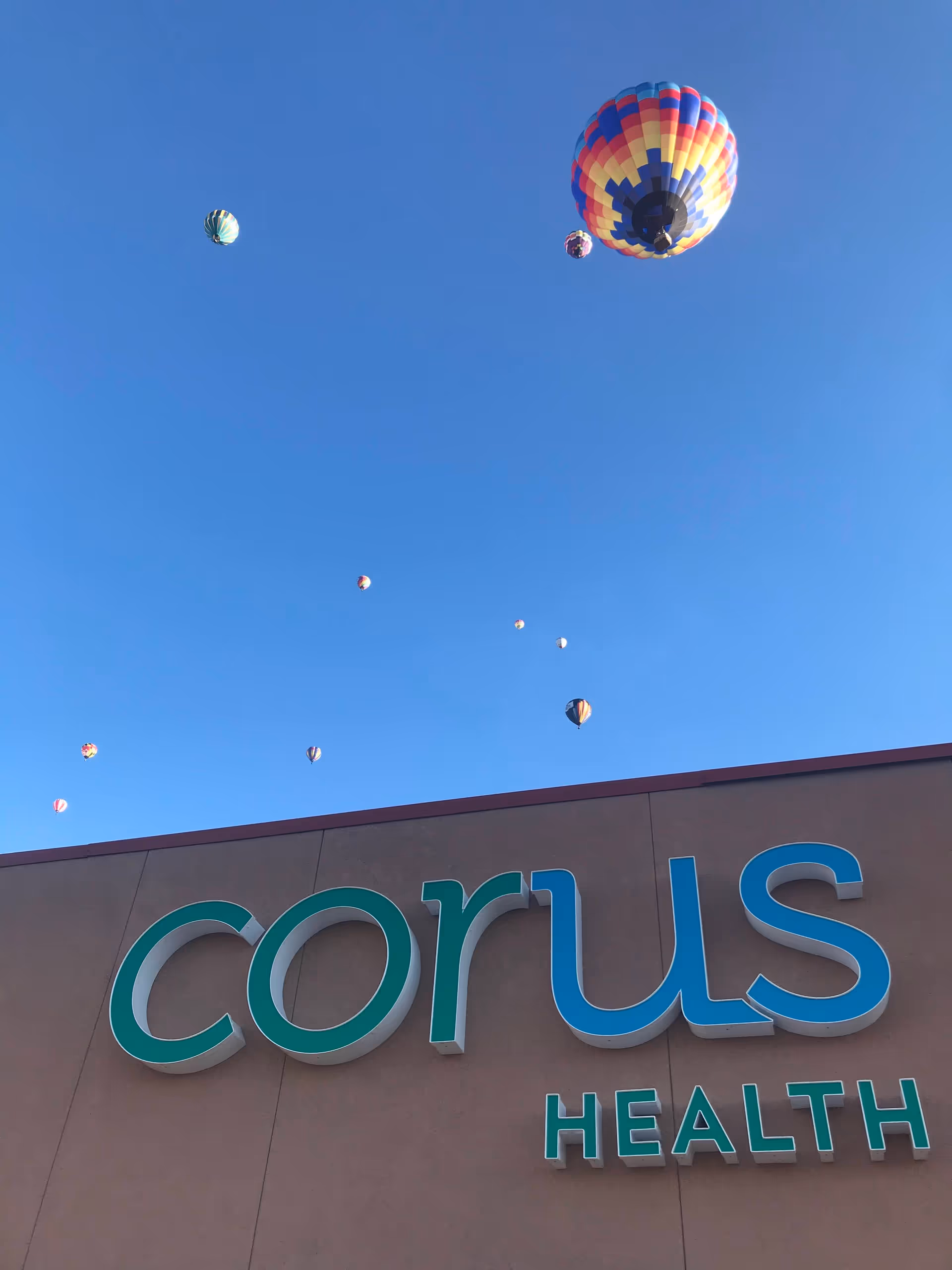 Blue sky with colorful hot air balloons above a Corus Health building sign.