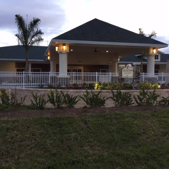 Front entrance of a single-story senior living facility with a covered porte-cochère, white railing, palm trees, and landscaping.