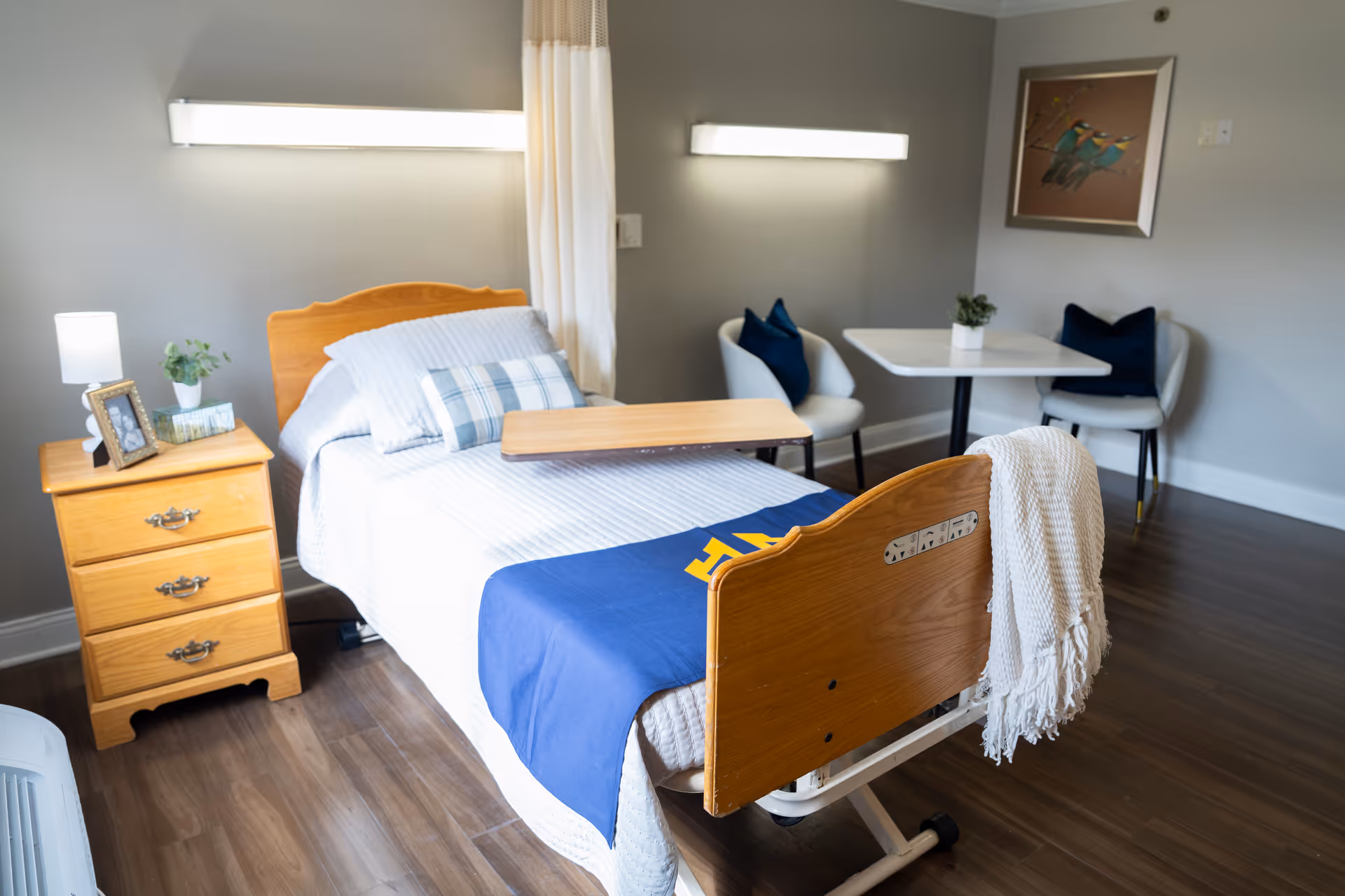 A neatly arranged bedroom in a healthcare facility featuring a single hospital bed with a blue blanket and white bedding. Next to the bed is a wooden nightstand with a lamp, a framed photo, a small plant, and a box of tissues. In the corner, there is a small white table with two white chairs, each with a dark blue cushion. A framed picture of birds hangs on the wall above the table. The room has wooden flooring and soft lighting.