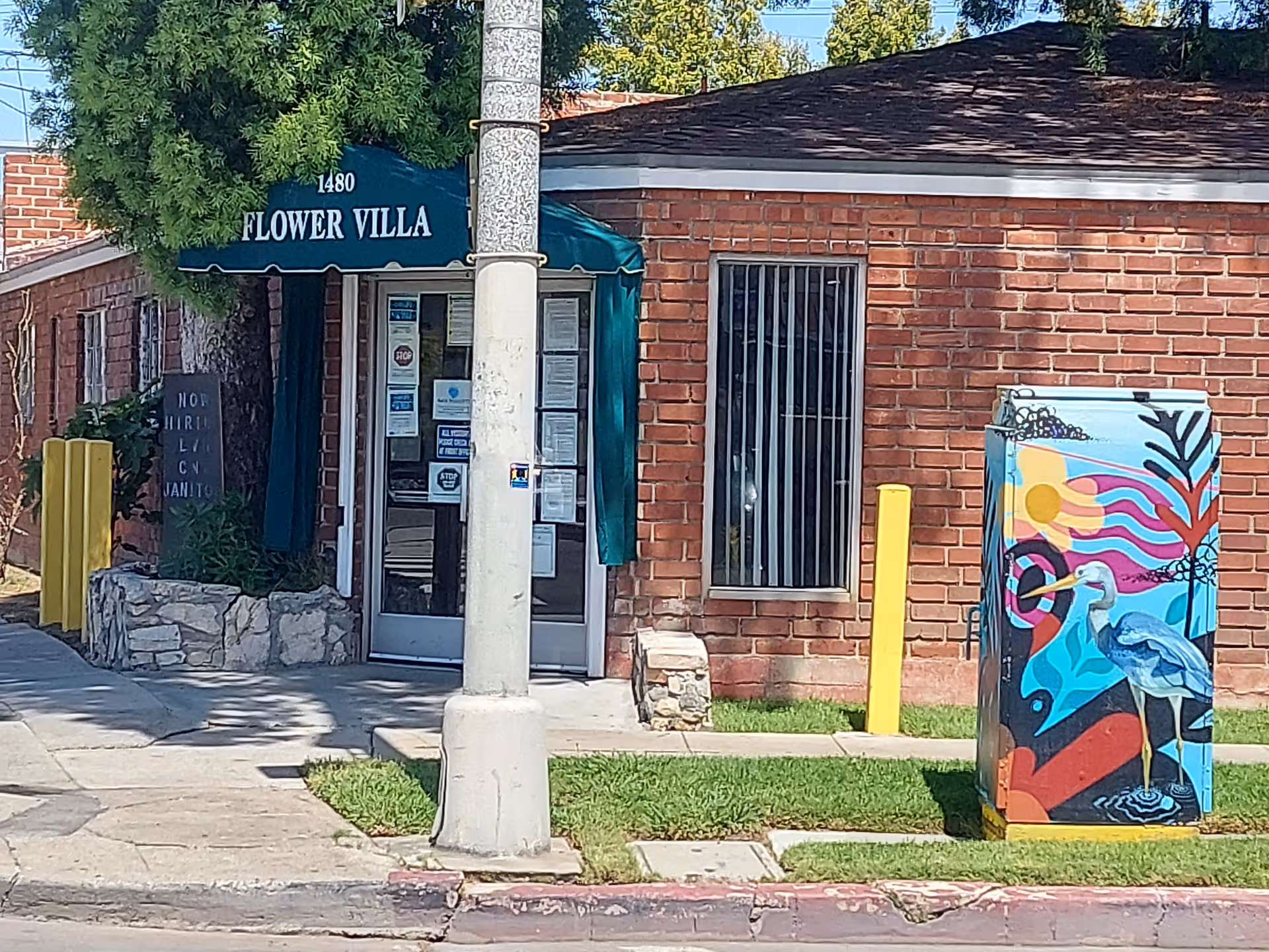 Brick exterior with Flower Villa entrance, green awning, metal door, and colorful utility box nearby.