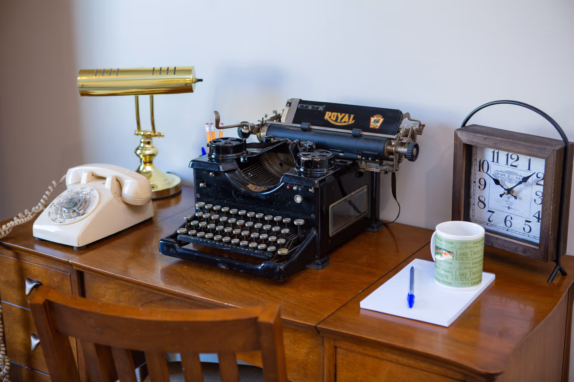 A wooden desk with a vintage black Royal typewriter, a cream-colored rotary dial telephone, a brass desk lamp, a wooden-framed clock showing 10:10, a white notepad with a blue pen, and a green and white coffee mug.