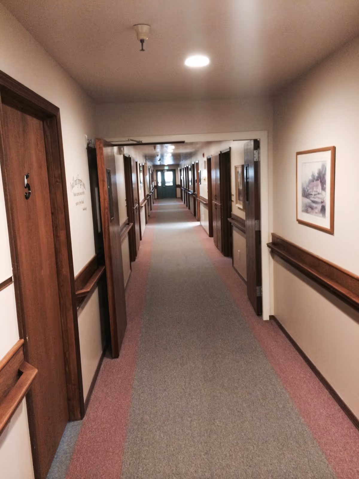 A long indoor hallway in a care facility with multiple wooden doors on both sides, beige walls, handrails, and framed artwork. The hallway is carpeted with a gray center and red borders, and a green exit door is visible at the far end.